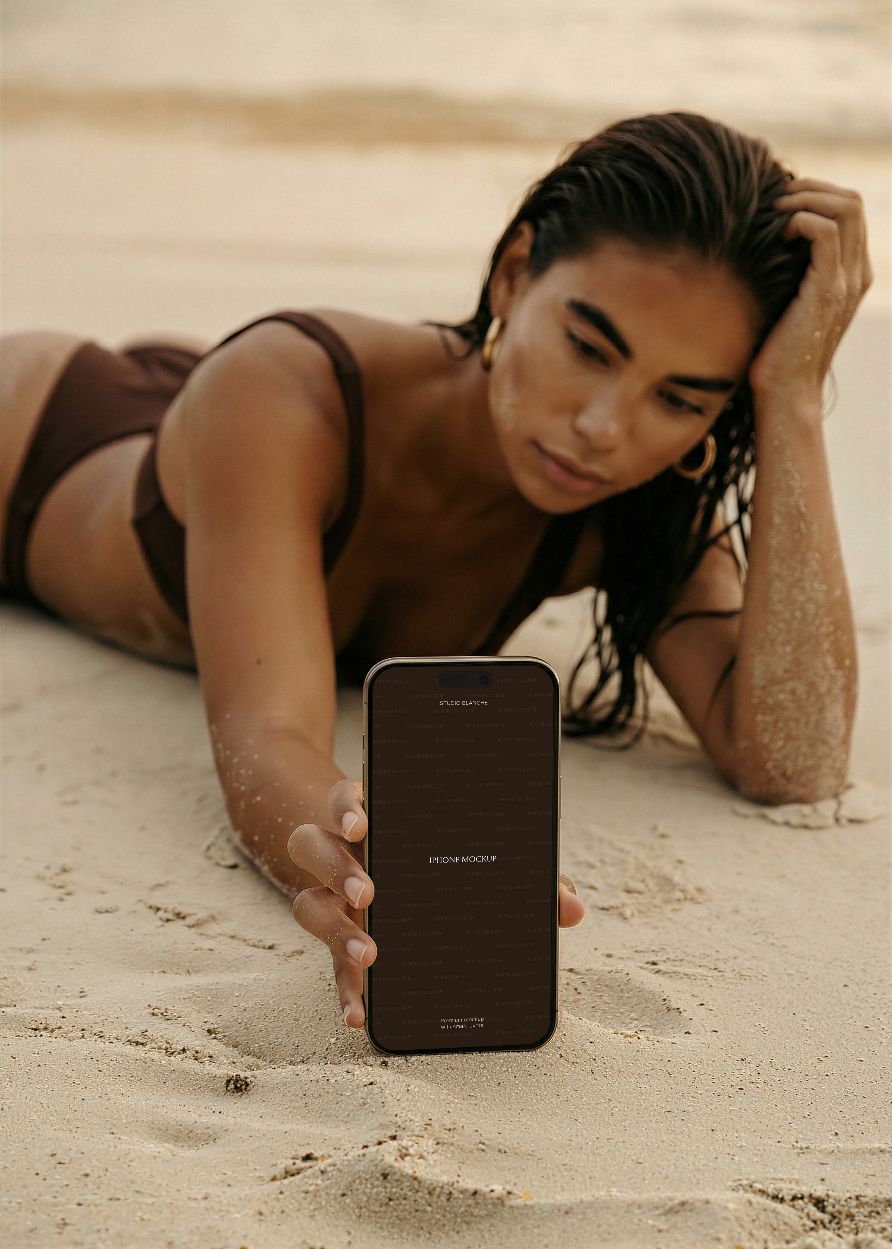 Beach iPhone mockup held by model on sand, dark screen placeholder, warm sun-toned lifestyle scene