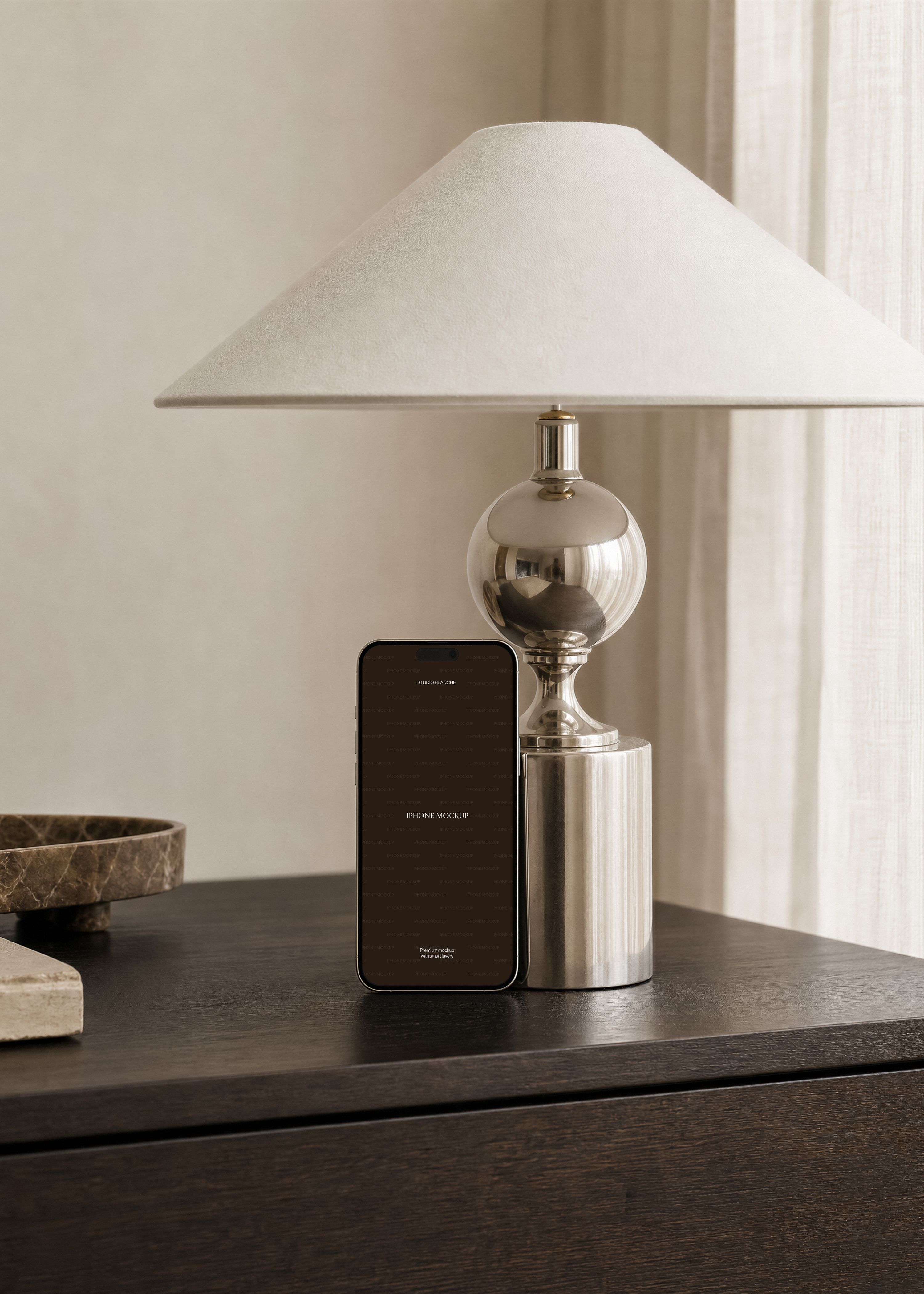 iPhone mockup leaning on chrome ball lamp on dark oak sideboard beside marble tray and travertine block, cream walls