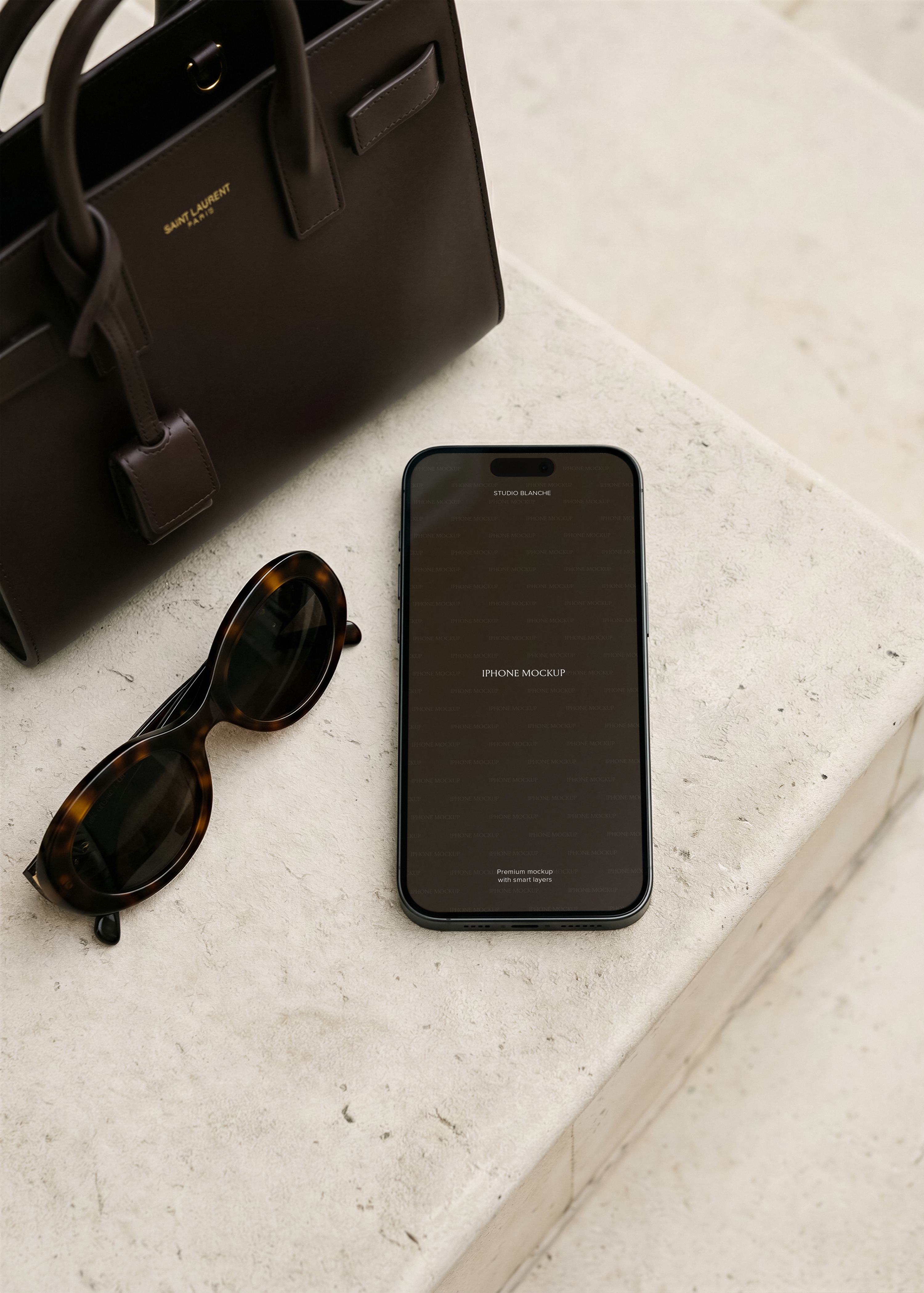 Dark iPhone with blank screen laid flat on pale stone surface beside tortoiseshell sunglasses and a dark structured leather tote, from Studio Blanche