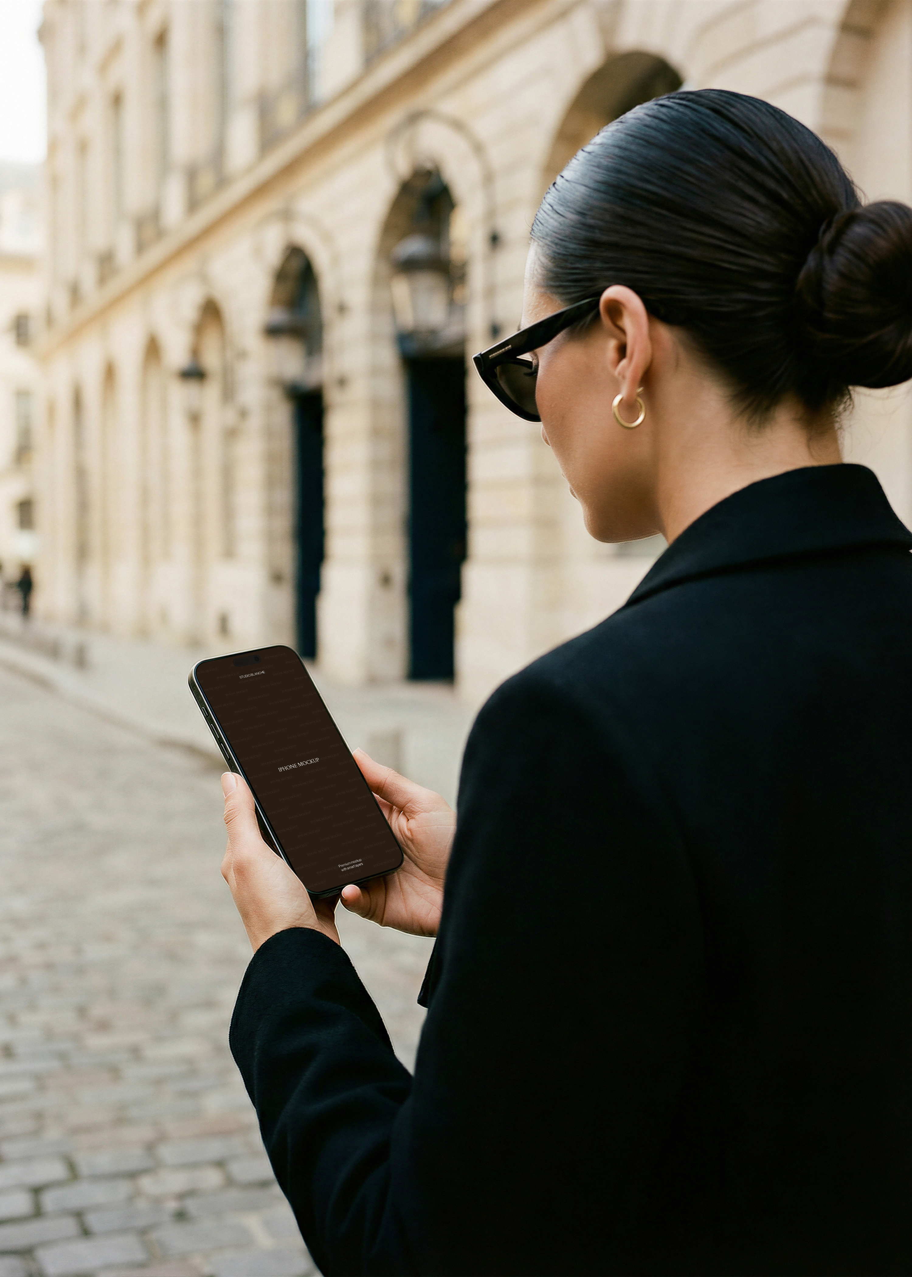 iPhone mockup held outdoors on Parisian cobblestone street, dark placeholder screen, Haussmann facade background