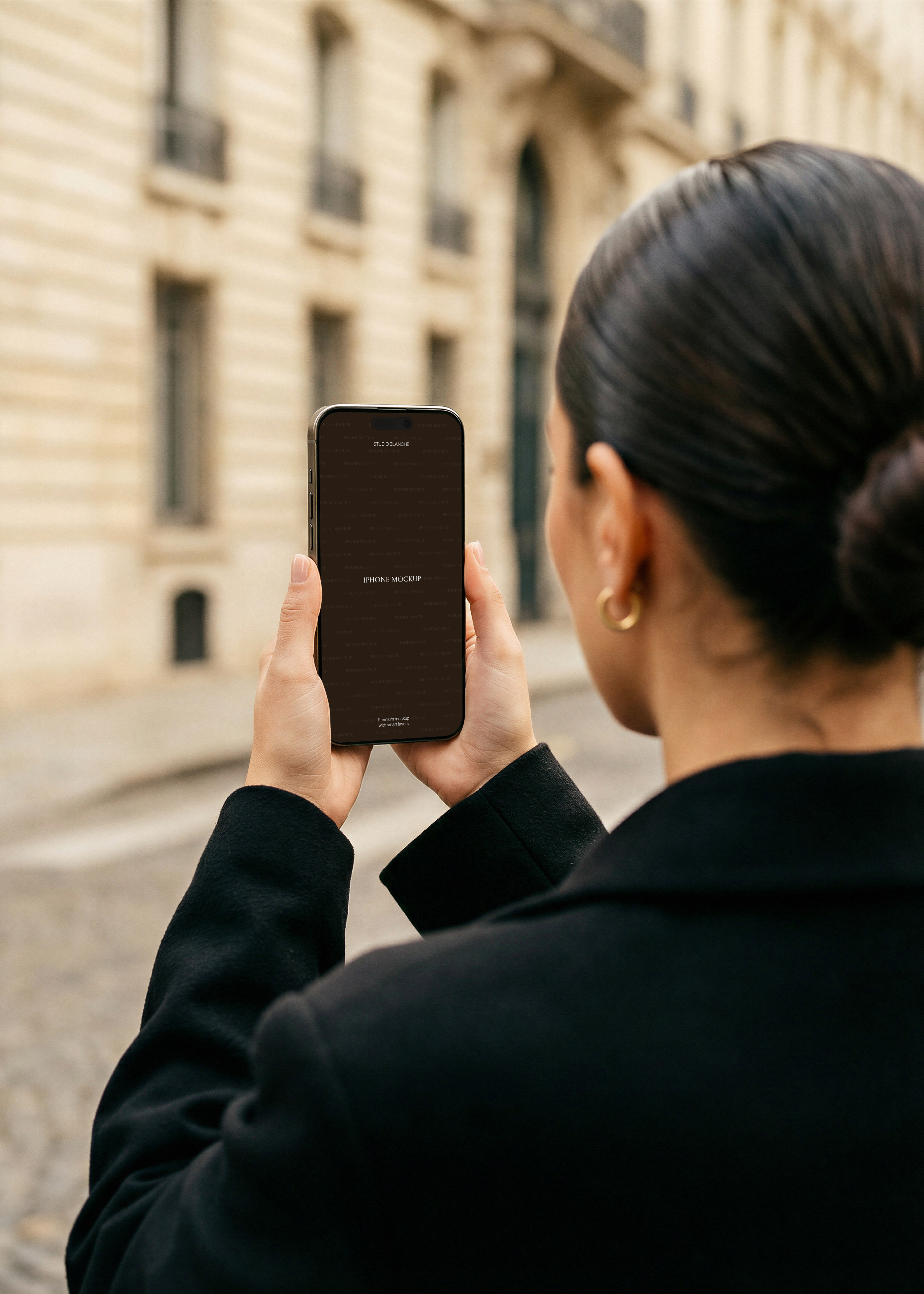 iPhone mockup held two-handed outdoors on Parisian street, dark placeholder screen, stone building behind