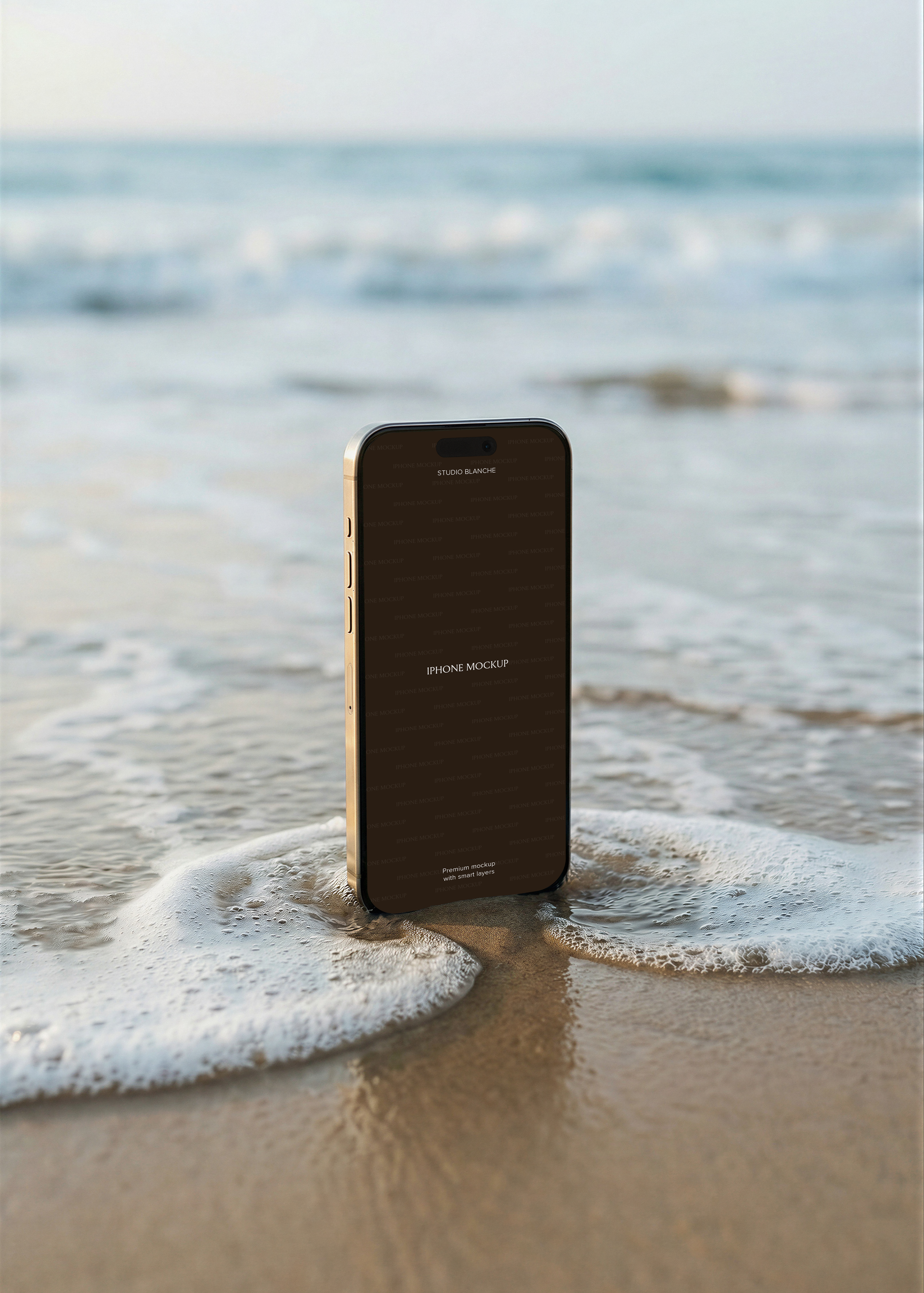 Gold iPhone standing upright in shallow wave on wet sand, open ocean and pale blue sky in soft focus background