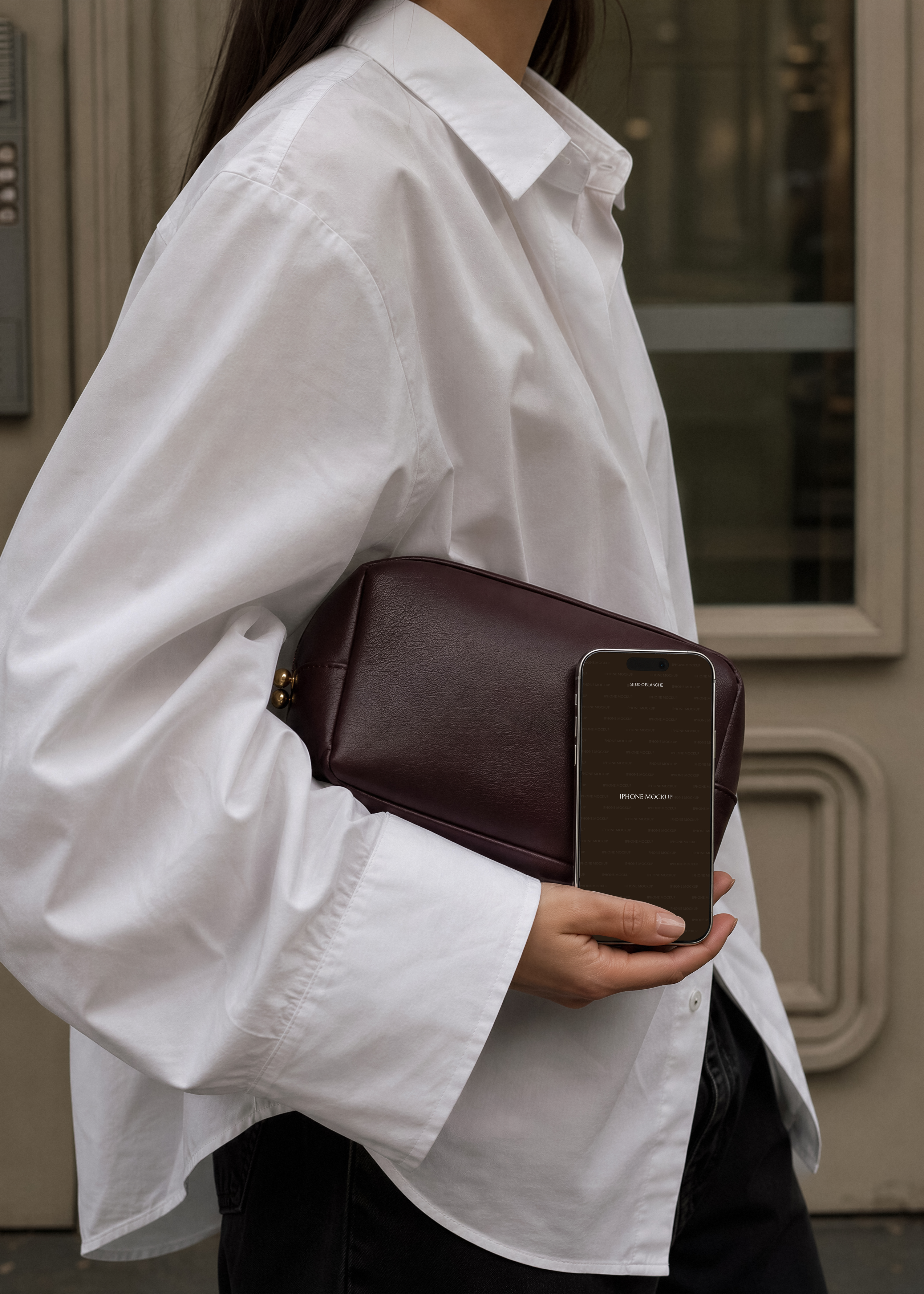 iPhone mockup with dark brown screen held against burgundy leather clutch, white oversized shirt, street doorway