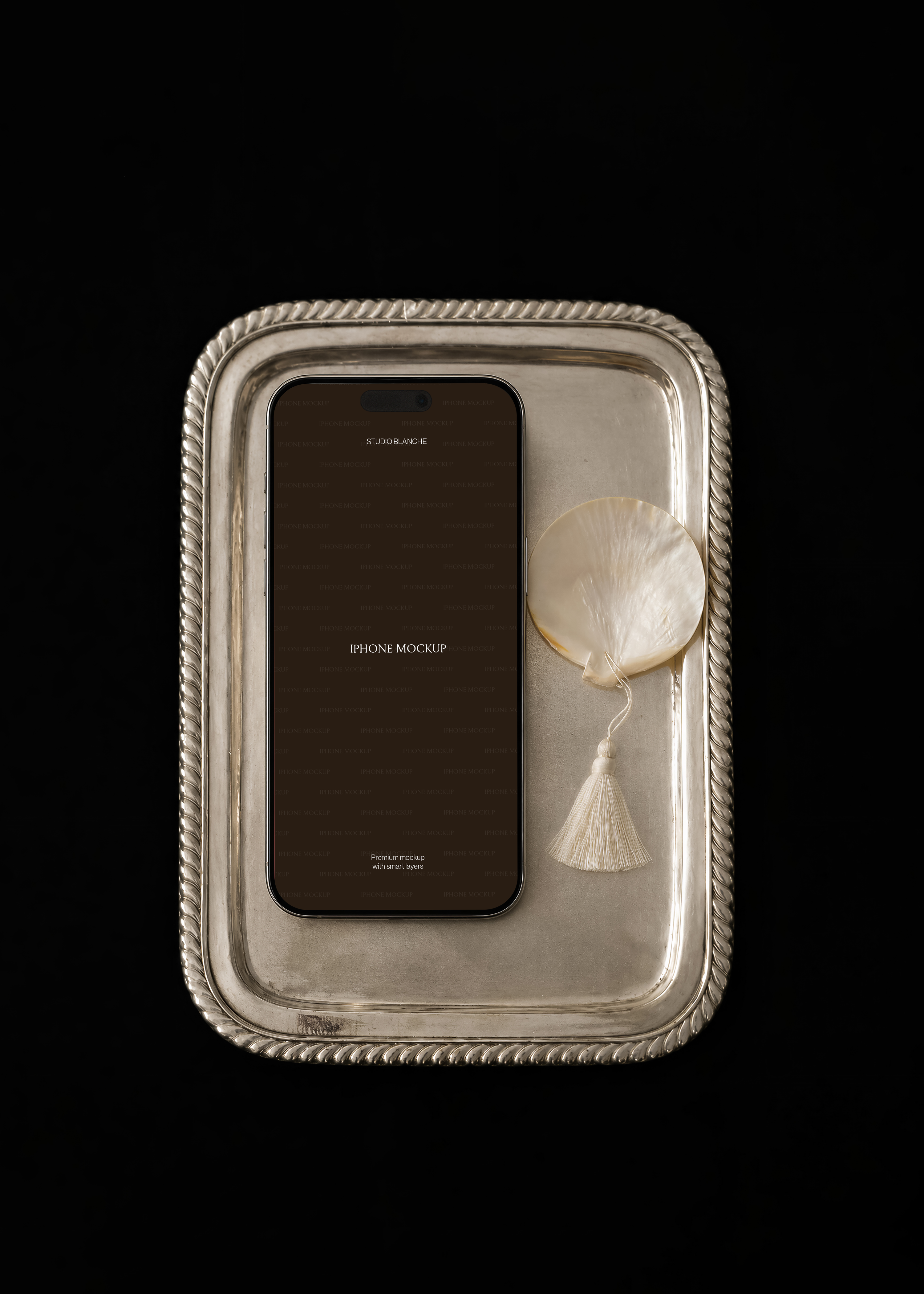 iPhone mockup with dark editable screen on rope-rimmed silver tray, pearl shell and cream tassel, dark editorial scene